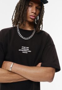 Czarny t-shirt z krótkim rękawem z białymi grafikami tekstowymi. Model nosi srebrny łańcuszek na szyi oraz srebrną bransoletkę.