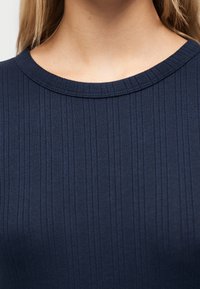 Κοντινό πλάνο του λαιμού και του άνω μέρους του στήθους ενός ατόμου που φοράει μπλε ναυτ ικό ριμπ πουκάμισο με στρογγυλή λαιμόκοψη, με μακριά ξανθά μαλλιά ορατά.