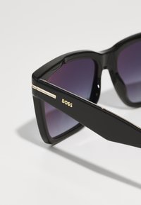 Óculos de sol retangulares pretos com lentes roxas em gradação. Apresentam acabamentos em ouro e o logótipo "BOSS" de um lado. Armação de plástico elegante.