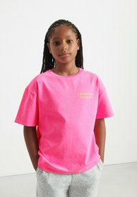 Różowy bawełniany t-shirt z krótkimi rękawami, na którym wydrukowano napis 'AMERICAN VINTAGE' w jaskrawożółtym kolorze. Połączony z szarymi spodniami dresowymi, stojący na neutralnym tle.