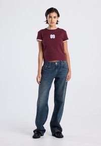 Maroon cropped t-paita, jossa on valkoinen kaulus ja '89' printti, yhdistettynä löysiin sinisiin farkkuihin ja mustiin korokepohjakenkiin neutraalilla taustalla.