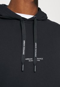 Felpa nera con cappuccio e lacci neri, con la stampa "Armani Exchange" e una piccola scritta bianca "Armani Exchange Milano New York" sul petto.