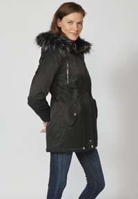 Črna parka jakna s kapuco iz umetnega krzna, zoženim pasom, žepi z zadrgo in sijočo teksturo. V paru z temno modrimi jeans hlačami.