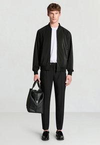 Giacca bomber nera con colletto a costine, abbinata a una camicia bianca, pantaloni neri e scarpe nere eleganti. Tiene una borsa in pelle nera.