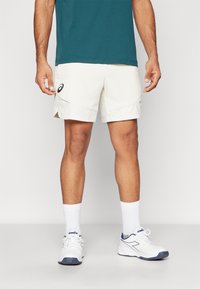 Männer Sportcreme-Shorts mit seitlichem Schlitz, kombiniert mit weißen Turnschuhen und Knöchelsocken. Mit einem Asics-Logo am Saum.