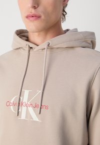 Felpa con cappuccio beige realizzata in tessuto morbido, con logo bianco e scritta rossa "Calvin Klein Jeans" sulla parte anteriore. Cordini regolabili.