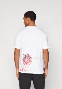 White t-shirt featuring a playful red graphic design on the back, with a character and yarn ball illustration. Short sleeves and crew neck.