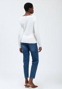 Weißes Langarmoberteil mit lockerer Passform, hat einen runden Rückenausschnitt und einen kleinen goldenen Akzent. Kombiniert mit blauen Jeans und braunen Plateausandalen.