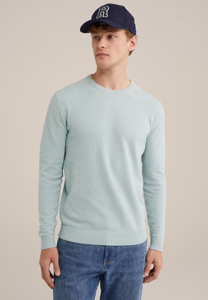 Pulover tricotat de culoare bleu deschis, cu un model texturat, guler rotund și manșete elasticizate. Asortat cu blugi de denim albastru închis și o șapcă bleumarin.