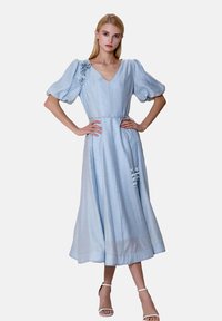 Robe bleu clair, longueur genoux, avec un décolleté en V, des manches bouffantes et un accent floral sur l'épaule gauche. Fabriquée en tissu texturé.