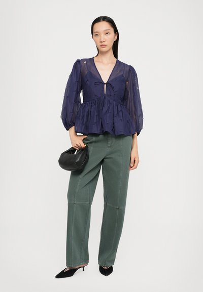 Blusa azul marino con peplum, mangas transparentes y detalle de lazo en el cuello, combinada con pantalones verdes de pierna ancha y un bolso negro.