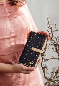 Black wallet with white polka dots, tan leather strap closure and gold hardware logo. Rectangular shape, smooth texture, compact design.