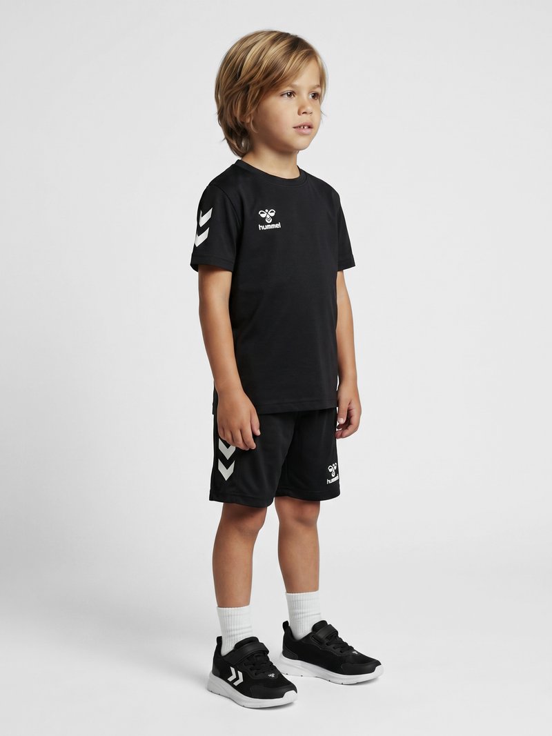 Ung dreng står iført sort sportstøj med hvide logoer, hvide sokker og sorte sneakers mod en ensfarvet lys baggrund.