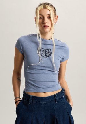 Młoda kobieta o jasnych blond włosach nosi dopasowany, lawendowy T-shirt z czarnym motywem serca oraz luźne, ciemne jeansy, trzymając ręce w kieszeniach.