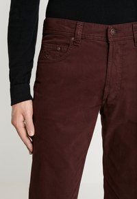 Pantaloni di cotone color marrone scuro con una texture liscia, dotati di una tasca frontale con dettaglio di cuciture e una chiusura tradizionale con bottone.