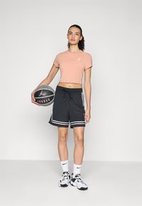 Lys rosa avkortet t-skjorte med en liten logo, svarte shorts med hvite striper, svart-hvite joggesko, holder en basketball.