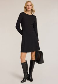 Schwarzes langärmeliges Kleid mit tailliertem Oberteil und ausgestellt A-Linien-Rock. Glatter Stoff, minimale Nähte, kombiniert mit knielangen schwarzen Stiefeln und einer Handtasche.