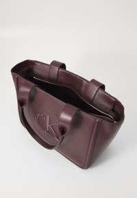 Sac cabas en cuir bordeaux avec deux poignées, fermeture éclair dorée et logo "CK" embossé sur le devant. Poche intérieure visible.