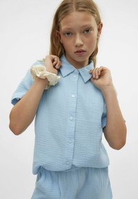 Gaiši zils īsais piedurknes krekls ar apkakles kakla izgriezumu, pogu aizdari un teksturētu audumu. Krēmkrāsas scrunchie ar krāsainiem punktiem ap plaukstu.