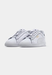 Bílé tenisky Puma se šněrováním, zlaté logo Puma na boku, černé logo Puma na jazyku, pohled zepředu na jednoduchém pozadí.