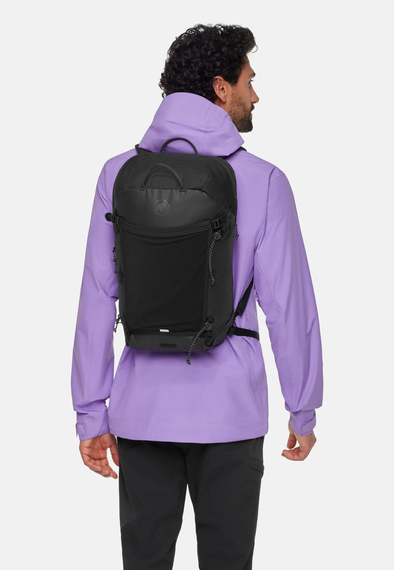Svart ryggsäck med en elegant design, tillverkad av slitstarkt material med dragkedjor, justerbara remmar och en logotyp på framsidan. Bärs av en person.