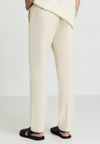 Beige Hosen im geraden Schnitt, mit glatter Textur und seitlichen Taschen. Getragen mit schwarzen Sandalen, von hinten betrachtet.