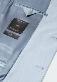 Intérieur d'un costume bleu clair montrant un tissu lisse et texturé avec des étiquettes noires marquées "MNG" et "SLIM FIT", indiquant les détails de taille et de matériau.