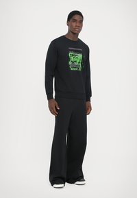 Schwarzer Sweatshirt mit grünem Grafikdruck, gerippten Bündchen und Rundhalsausschnitt. Kombiniert mit locker sitzenden schwarzen Hosen und schwarzen Sneakers.