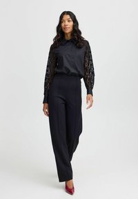 Schwarze Bluse mit Spitzen-Langärmeln, kombiniert mit hochtaillierten schwarzen Hosen und roten spitzen High Heels. Glatte Textur, maßgeschneiderte Passform.