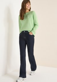 Jasnozielona bluza z nadrukowanym logo o fakturze, w zestawieniu z ciemnoniebieskimi rozszerzanymi jeansami z kontrastowym przeszyciem oraz srebrnymi butami sportowymi.