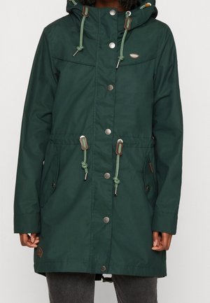 Manteau à capuche vert foncé avec boutons-pression à l'avant, cordons réglables à la capuche et à la taille, poches latérales, porté sur un pantalon gris.