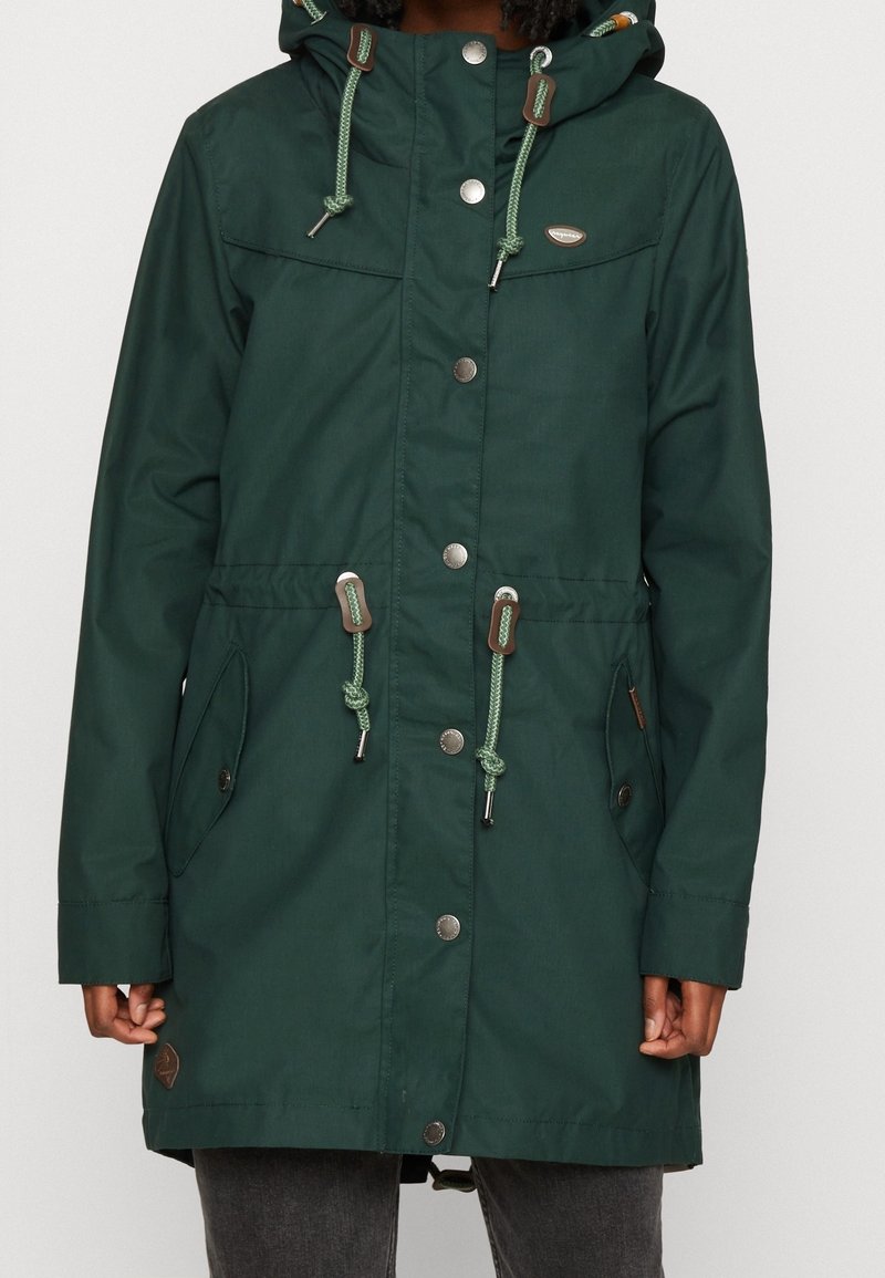 Manteau à capuche vert foncé avec boutons-pression à l'avant, cordons réglables à la capuche et à la taille, poches latérales, porté sur un pantalon gris.