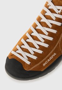 Hnedé semišové turistické topánky s bielymi šnúrkami a čiernym gumovým prstom, s logom Scarpa na bočnej strane.
