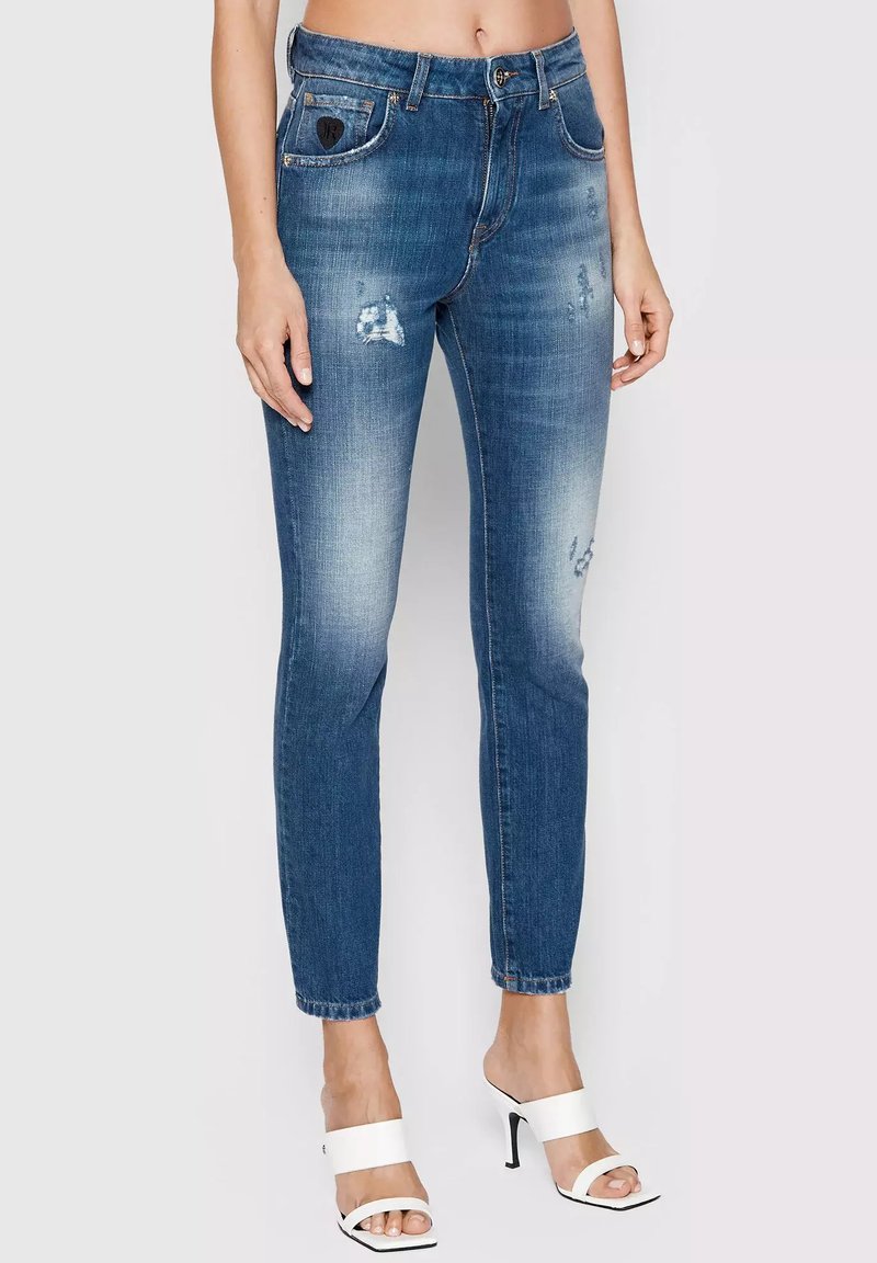 Donna che indossa jeans skinny strappati blu e sandali con tacco bianchi, in piedi contro uno sfondo chiaro e uniforme.