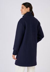Cappotto oversize blu navy con collo alto, polsini a coste e tessuto strutturato. Presenta una linea dritta e spacchi laterali per facilitare i movimenti.