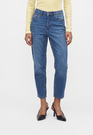 Kvinde iført blå højhældede jeans med lige ben, lysegul langærmet top og sorte spidse højhælede slingback-sko.