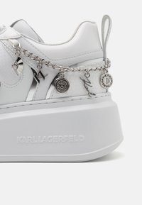 Sneaker à plateforme blanche avec chaîne argentée et multiples breloques en métal, avec un logo Karl Lagerfeld sur la semelle.
