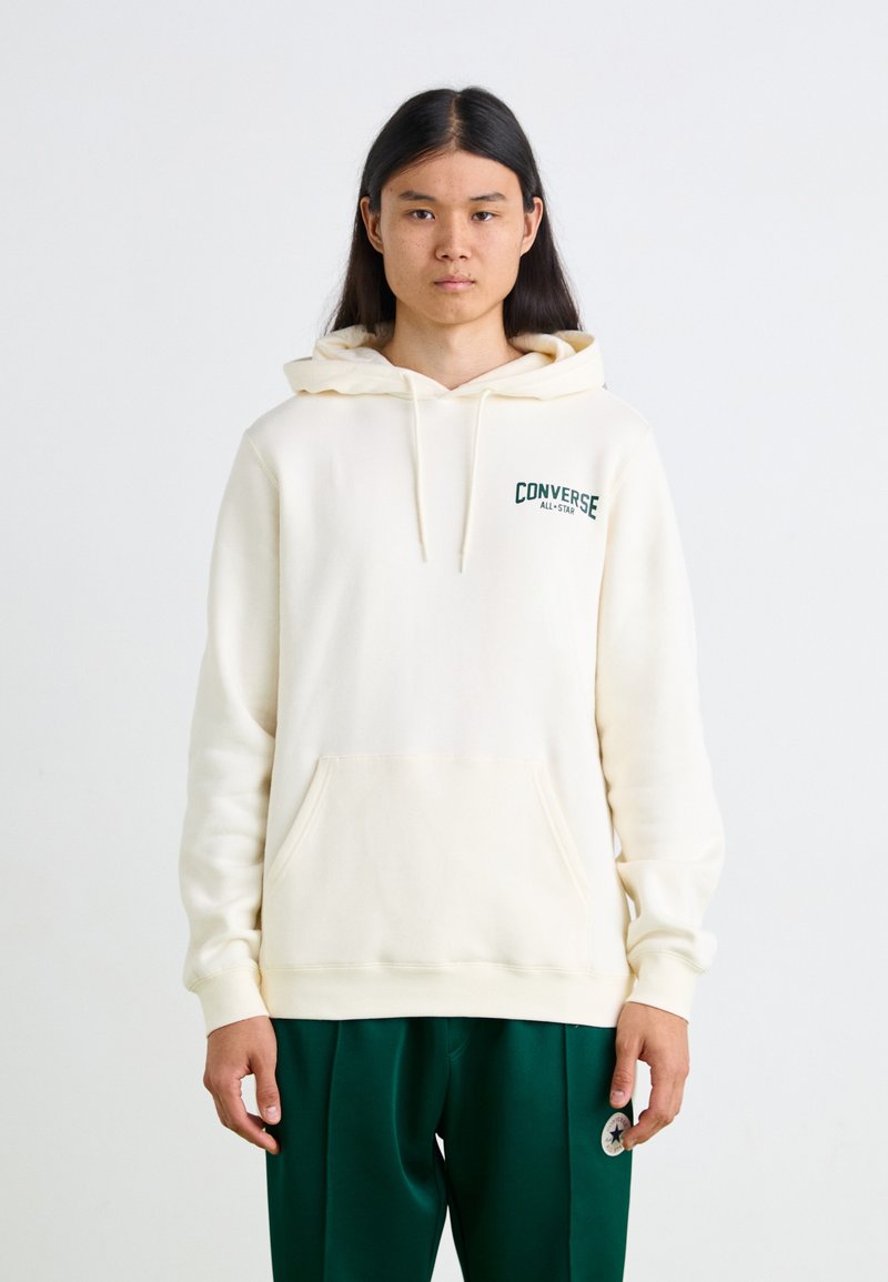 Sudadera crema hecha de un tejido suave con un bolsillo delantero. Presenta un logo verde de "Converse All-Star" en el lado izquierdo del pecho. Corte holgado.