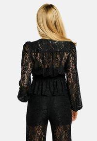 Melns mežģīņu tops ar gariem pūderveida piedurknēm, kas rotāts ar volāniem apakšmalā un ziedu rakstu. Teksturētā auduma kārtas ir caurspīdīgas.