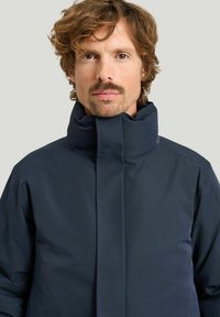 Marinblå ytjacka med hög krage, tillverkad av ett slätt, vattenavvisande tyg. Har en elegant, minimalistisk design och dolda stängningar.