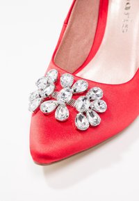 Chaussure plate en satin rouge à bout pointu avec un groupe de grandes pierres précieuses transparentes sur le devant.