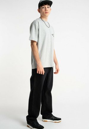 Młody mężczyzna w jasnoszarej, oversize'owej koszulce, czarnych szerokich spodniach, czarno-białych trampkach, z łańcuszkiem na szyi i czarną czapką, stoi.