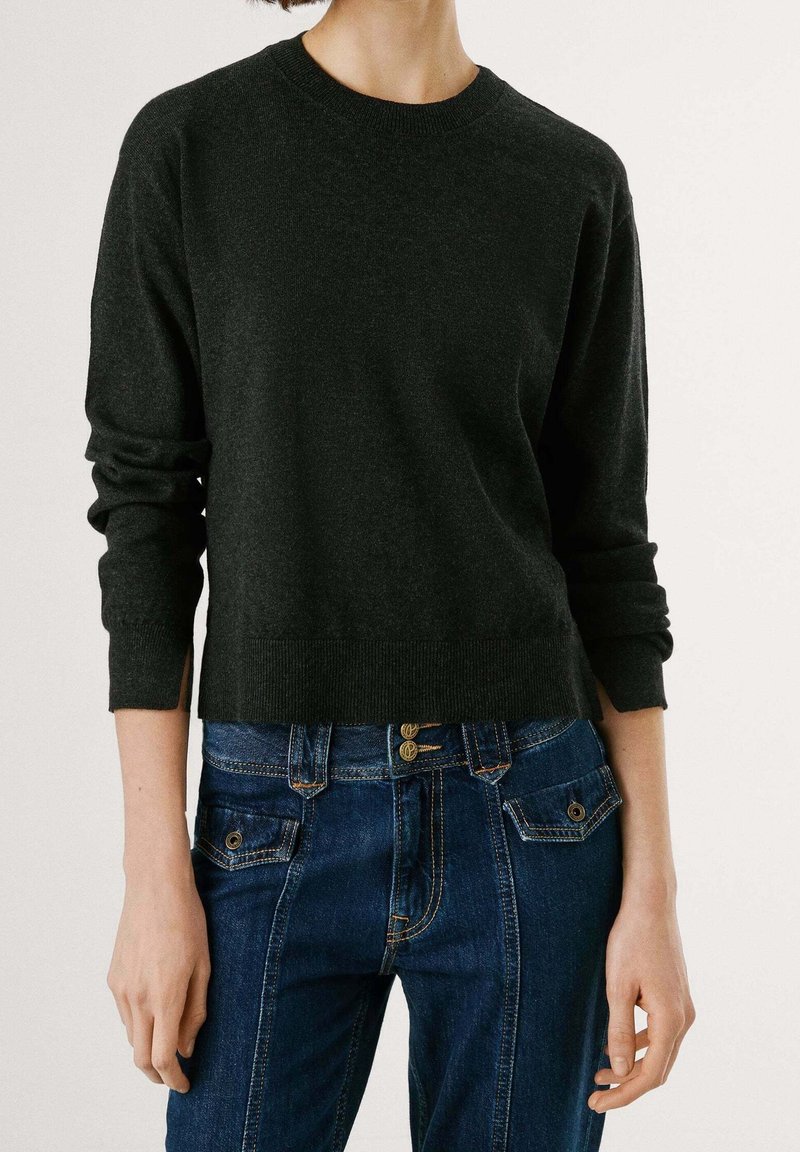 Femme portant un pull en laine gris foncé à manches longues associé à un jean en denim bleu taille haute avec des poches à boutons.