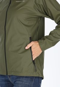 Giacca impermeabile verde oliva con zip frontale e tasche laterali; presenta dettagli neri sulla zip e una texture liscia e lucida.