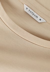 Beige pamut póló lekerekített nyakrésszel és a "TNTUUM" márkanevével valamint "M" mérettel ellátott címkével, sima szövettextúrával.