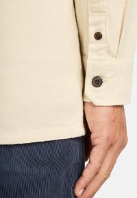 Crèmekleurig shirt met een textuurafwerking, voorzien van een knoopmanchet in donker hout, met ribbelstof die eronder zichtbaar is.