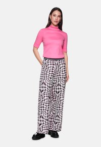 Woman standing with hands in pockets wearing a pink short-sleeve turtleneck and wide-leg patterned pants with black shoes.