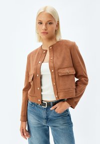Bruine faux suède cropped jas met een ronde hals, knoopsluiting, twee voorkruiszakken en een gladde textuur, gecombineerd met een witte top en blauwe jeans.