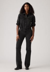 Sort denimskjorte med forlommer og hvid stikning, parret med sorte flared jeans og sorte støvler. Modellen står foran en neutral baggrund.
