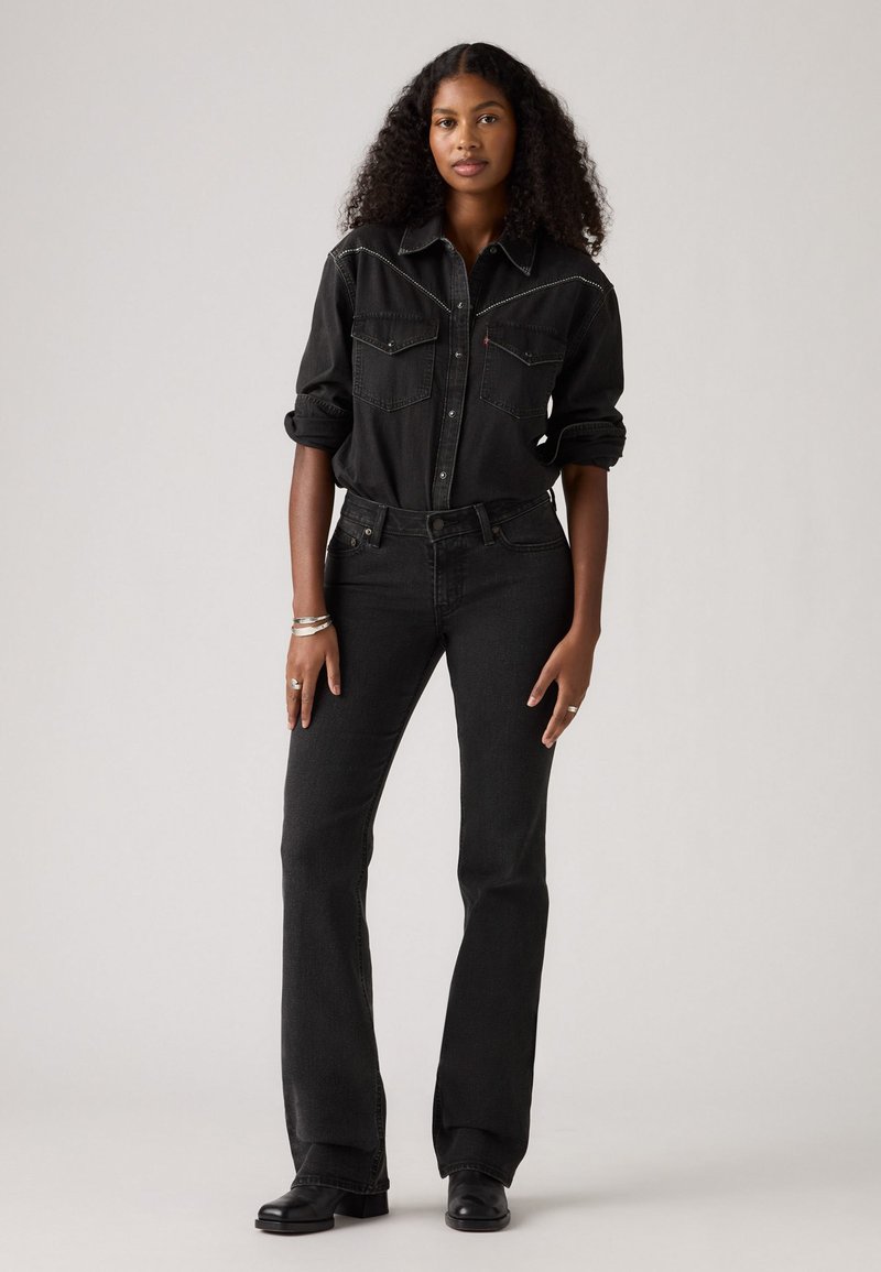 Sort denimskjorte med forlommer og hvid stikning, parret med sorte flared jeans og sorte støvler. Modellen står foran en neutral baggrund.